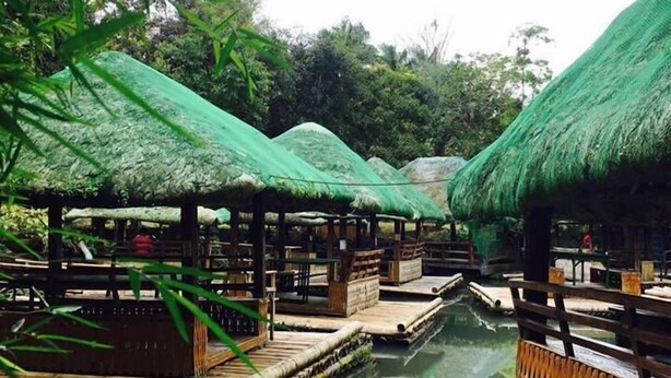 A bamboo raft with thatched roofs floating on a calm body of water, surrounded by lush greenery.