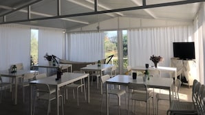 A spacious indoor area with multiple tables and chairs, large windows with curtains, and a television on a table. - Giota Rooms (Pineios)