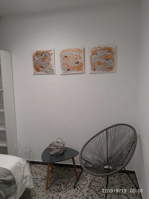 Una habitación con una cama, una mesita con una cesta, una silla y tres cuadros abstractos en la pared.