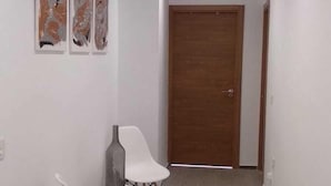 A hallway with a wooden door at the end, white walls, and three abstract paintings on the left wall. - Residencial Fontanillo (Las Palmas de Gran Canaria)