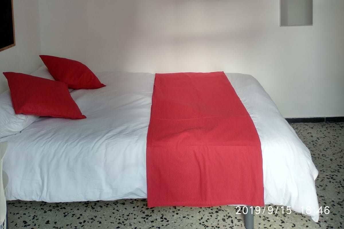 Un dormitorio con una cama, almohadas rojas, una colcha blanca y un edredón rojo. En la pared, hay un cuadro de un coche rojo y una planta en maceta.