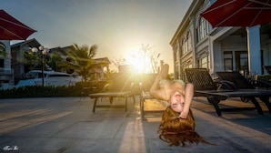 A poolside area with lounge chairs and umbrellas, a building with classical architecture, and a sunset in the background. - Zan- Newlife Halong Apartment (Bai Chay)