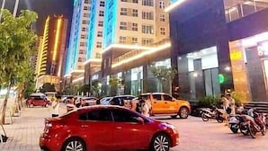 A high-rise building with multiple floors, illuminated by blue and white lights, and surrounded by parked cars and motorcycles. - Zan- Newlife Halong Apartment (Bai Chay)