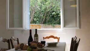 Ein Esstisch mit Blick auf Bäume durch ein Fenster, eine Weinflasche, ein Weinglas, Brot, Käse und eine Obstschale.