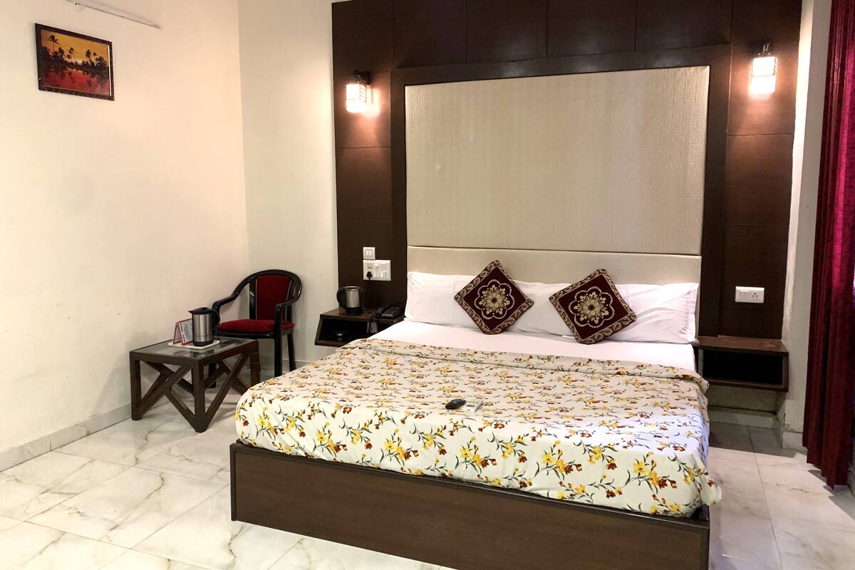 Quarto com cama grande, uma cadeira, mesa pequena e luminária na parede.