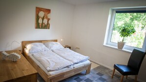 A bedroom with a wooden bed, a desk, a chair, a window with a plant, and a painting on the wall. - High quality and fully equipped apartment only 3km from the Baltic Sea (Stakendorf)