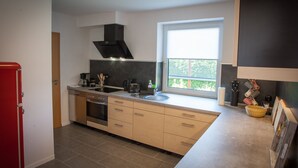 A modern kitchen with a built-in oven, a stovetop, a black range hood, a window with a view, and a red refrigerator. - High quality and fully equipped apartment only 3km from the Baltic Sea (Stakendorf)