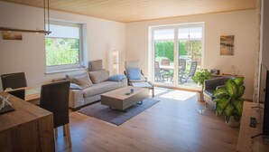 A spacious living room with a wooden floor, a sofa, a coffee table, and a dining area. There is a sliding glass door leading to an outdoor patio with a table and chairs. - High quality and fully equipped apartment only 3km from the Baltic Sea (Stakendorf)