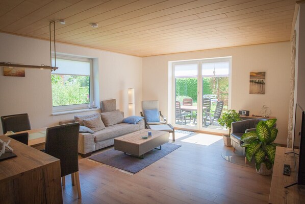 A spacious living room with a wooden floor, a sofa, a coffee table, and a dining area. There is a sliding glass door leading to an outdoor patio with a table and chairs.