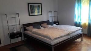 A bedroom with a large bed, two bedside tables, a nightstand, a framed picture on the wall, and a window with curtains. - Garden view in the Palatinate Forest - 80 m² with 3 bedrooms & terrace (Bundenthal)