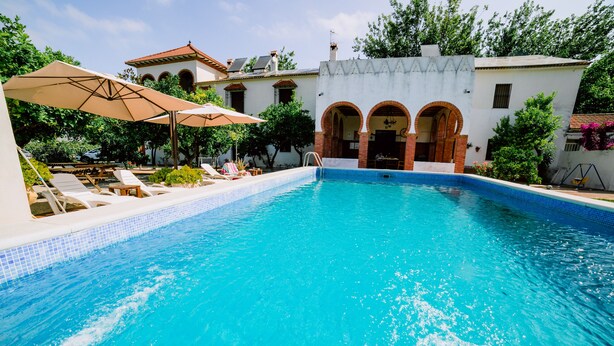 Una piscina con sillones de descanso y sombrillas, rodeada de edificios con arcos y un cielo despejado.