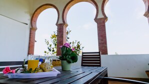Una mesa con alimentos para el desayuno, una planta en maceta y un banco en un espacio al aire libre cubierto con arcos.