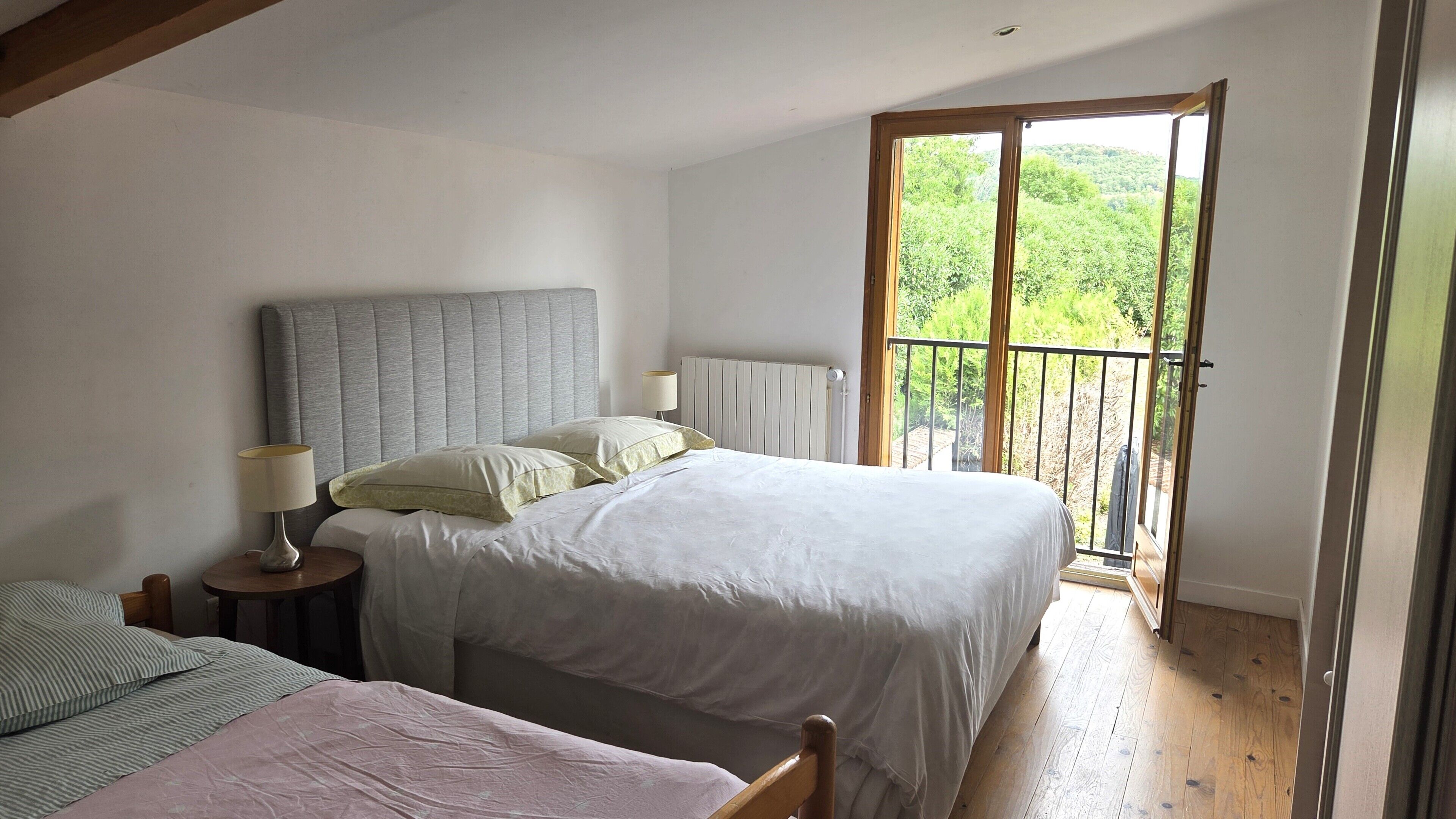 Un dormitorio con cama, mesita de noche, lámpara y vista a la vegetación a través de un balcón.