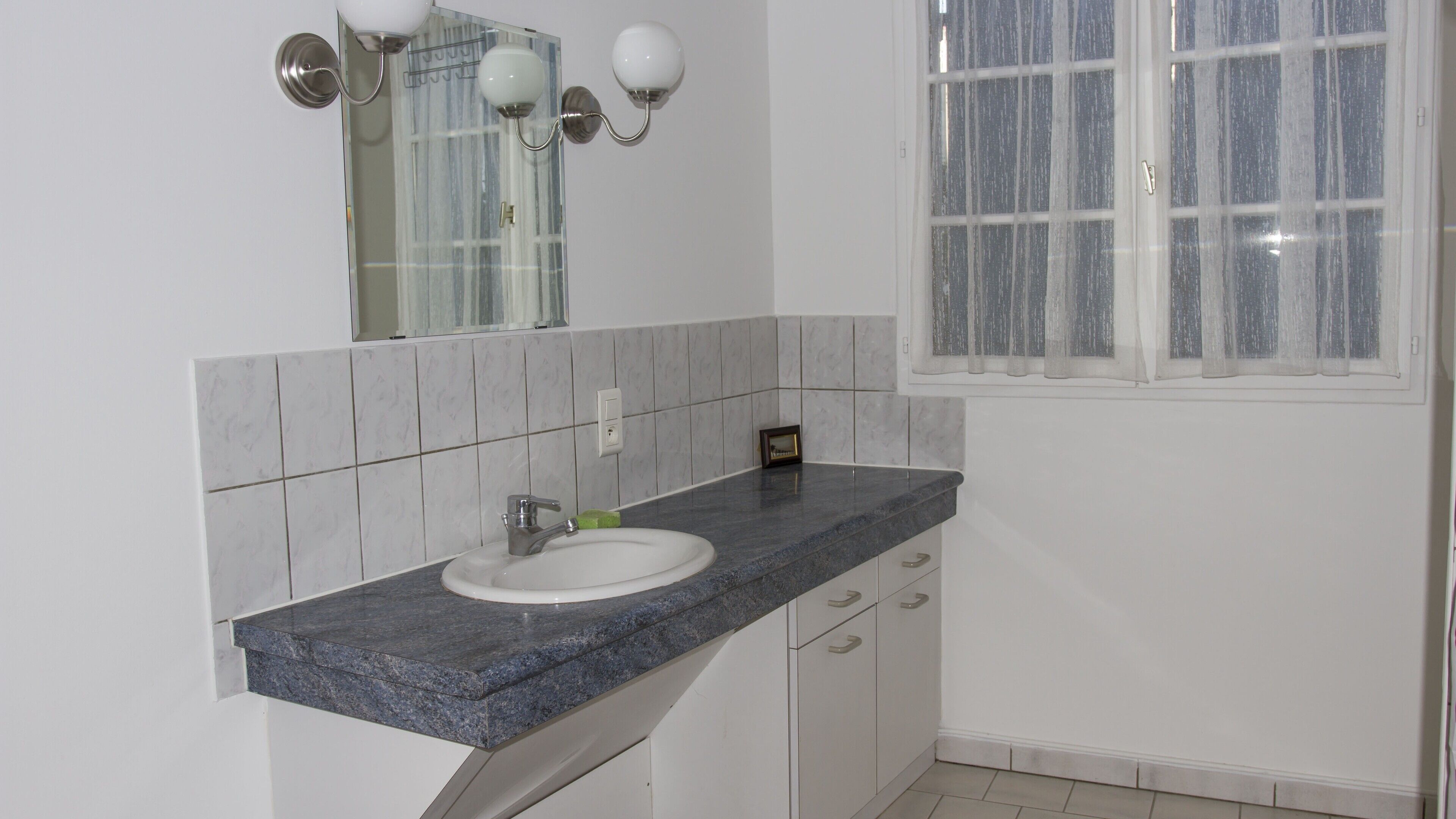 A bathroom with a white sink, granite countertop, and a window with a white curtain.