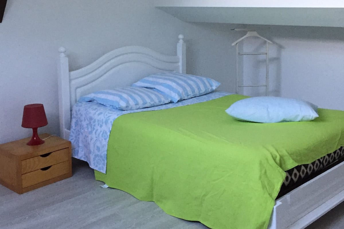 A single bed with a green bedspread and white pillows, a wooden nightstand with a red lamp, and a white wardrobe.