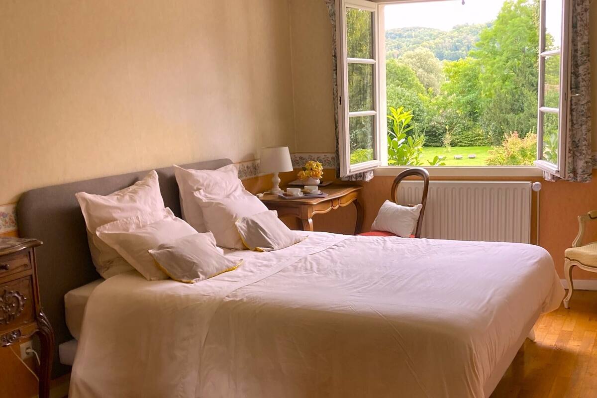 A neatly made bed with white linens, a wooden nightstand with a lamp, a chair, and a window with a view of greenery.