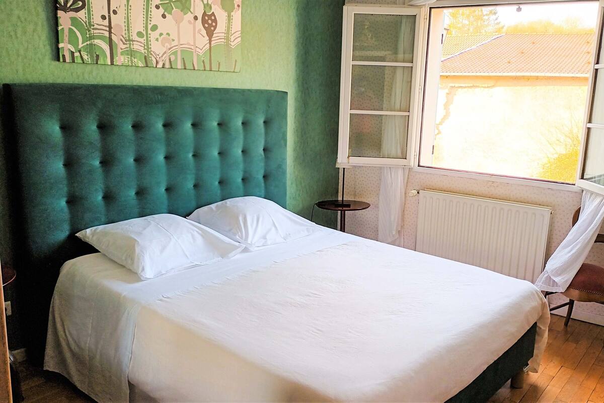 A bed with white linens, a teal upholstered headboard, and a window with a view of a house.
