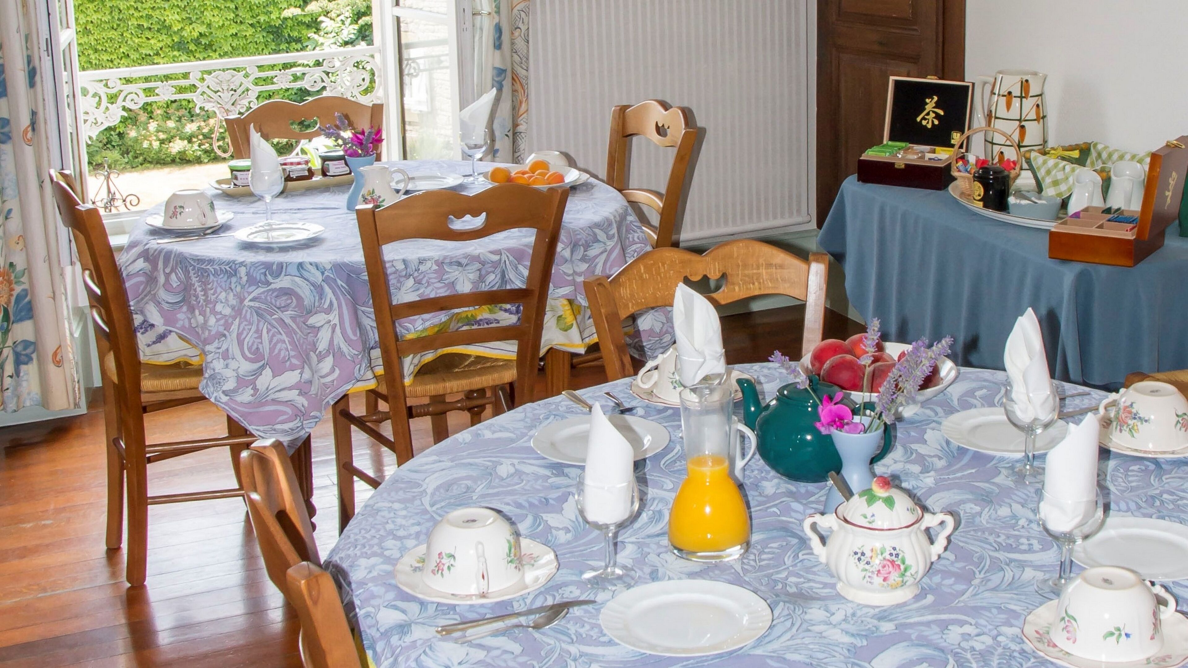 Een ontbijtruimte met ronde tafels, gedekt voor het ontbijt, met bloemenlinnen, wit porselein servies en diverse ontbijtartikelen.