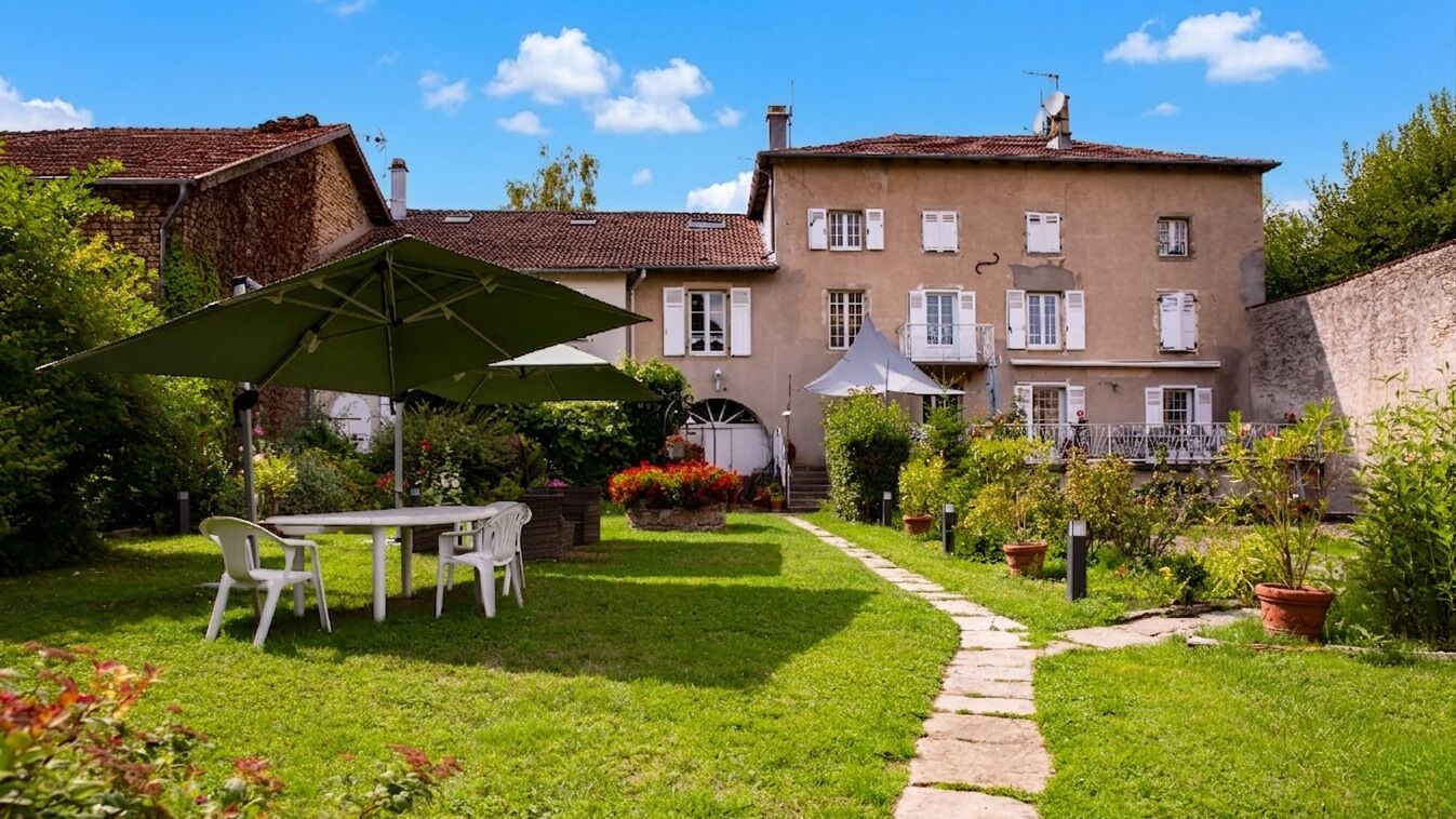 A courtyard with a table and chairs, surrounded by a garden and a building with a tiled roof.