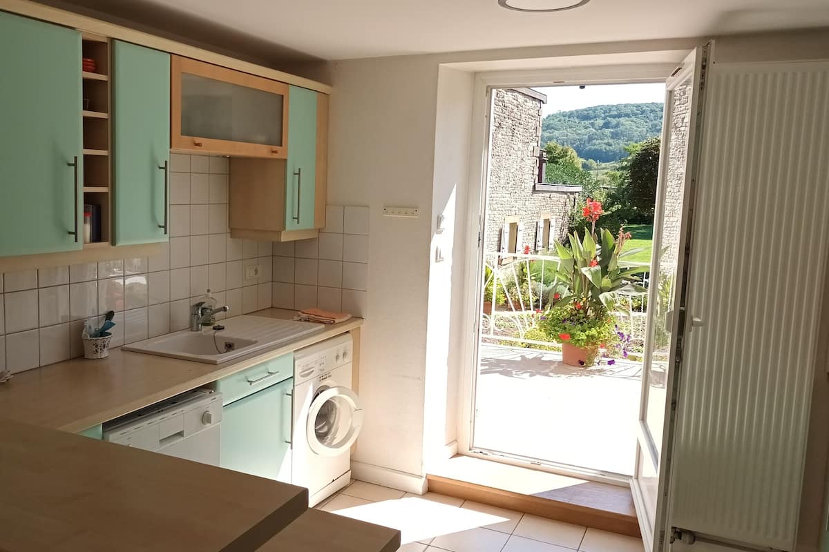 A kitchen with a washing machine, a coffee maker, and a view of a garden through an open door.