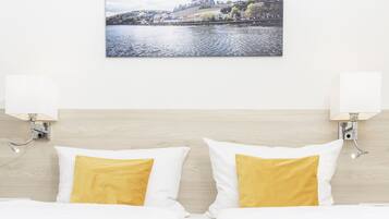 A hotel room with a bed, two white pillows with yellow accents, a wall-mounted picture of a landscape, and two wall-mounted lamps.