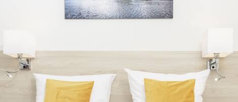 A hotel room with a bed, two white pillows with yellow accents, a wall-mounted picture of a landscape, and two wall-mounted lamps.