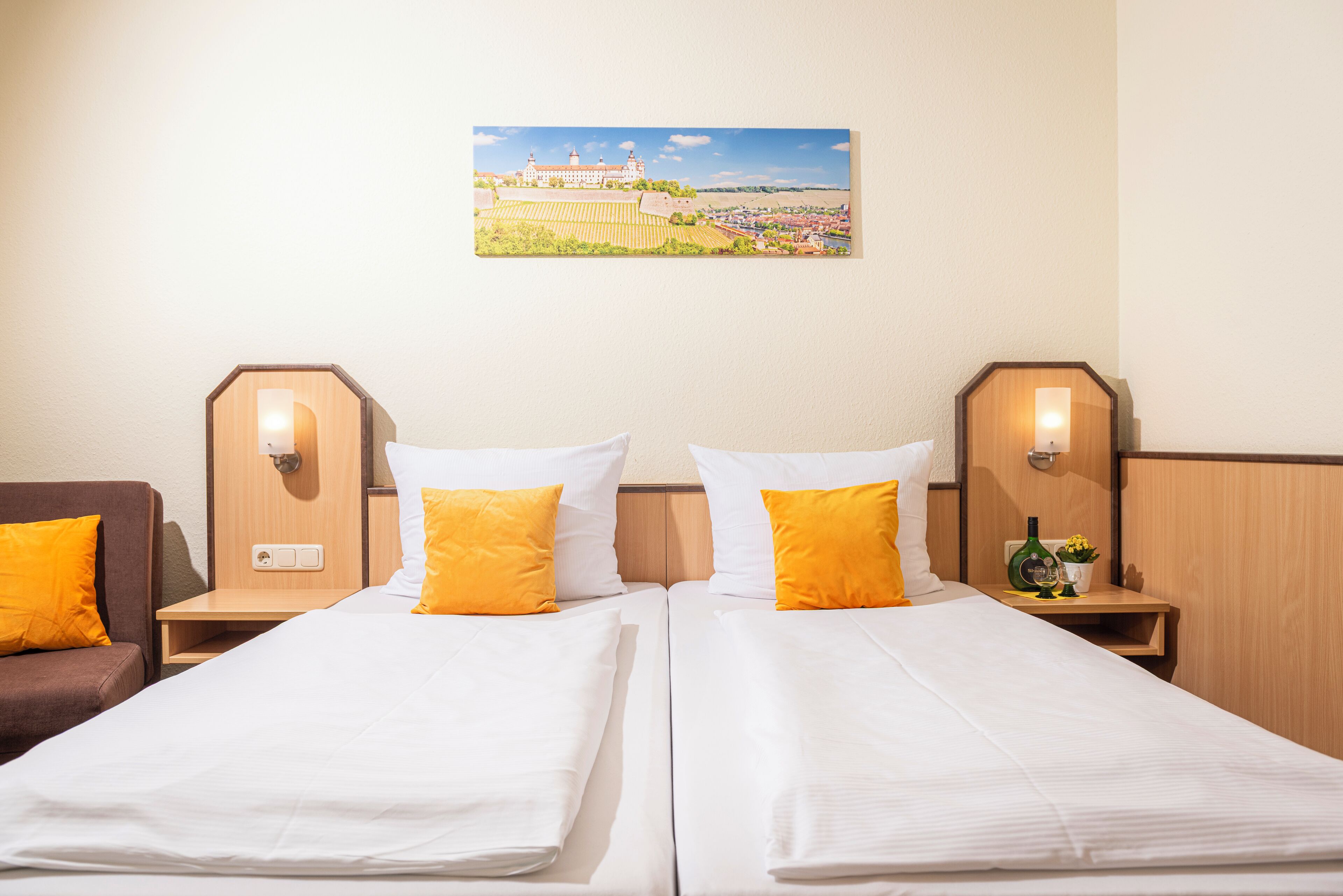 A hotel room with two beds, each with white linens and orange pillows, a painting of a castle on the wall, and wooden headboards with wall sconces.