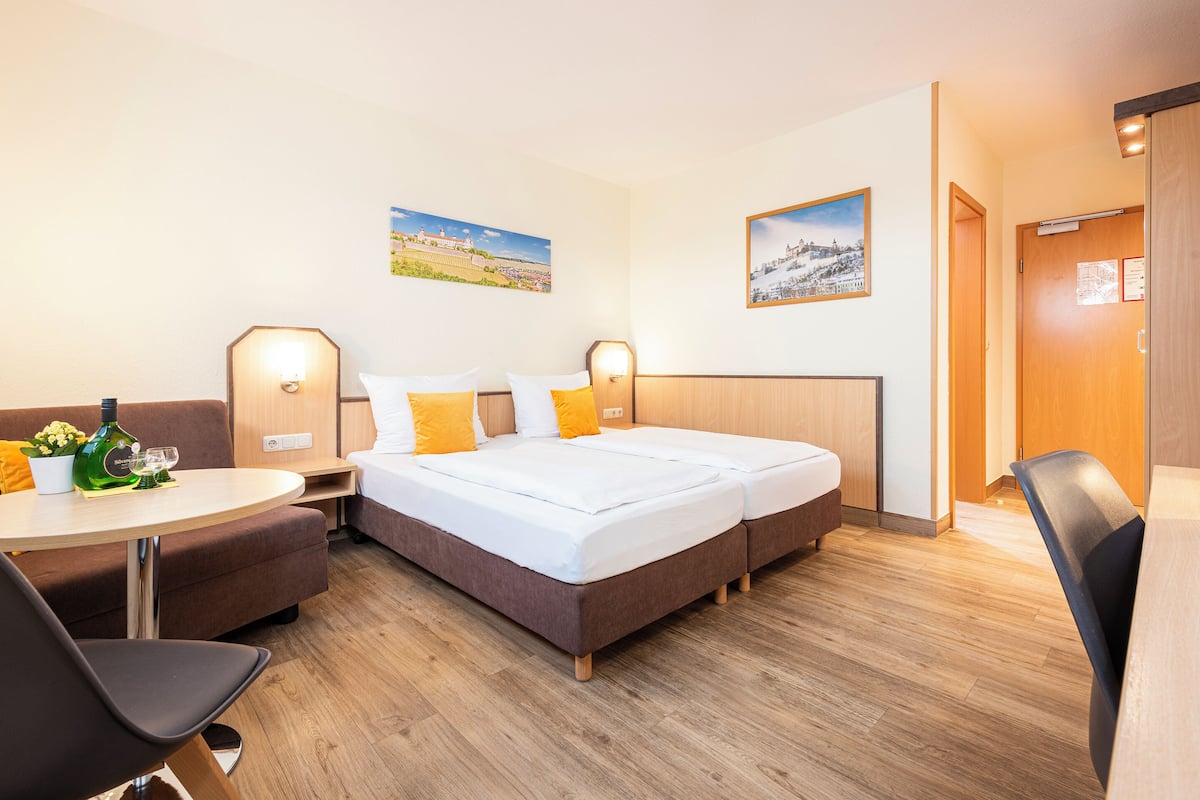 A hotel room with a double bed, a sofa, a small table with a bottle and glasses, and two wall-mounted lamps.