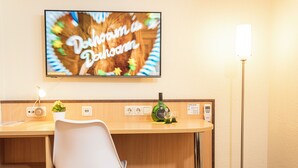 Hotellihuone, jossa on puinen työpöytä, valkoinen tuoli, televisiossa näkyy viesti, huoneessa on pullo alkoholia ja yöpöydällä lamppu.