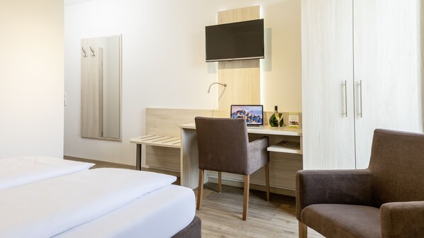 A hotel room with a bed, a desk with a computer, a chair, and a TV mounted on the wall.