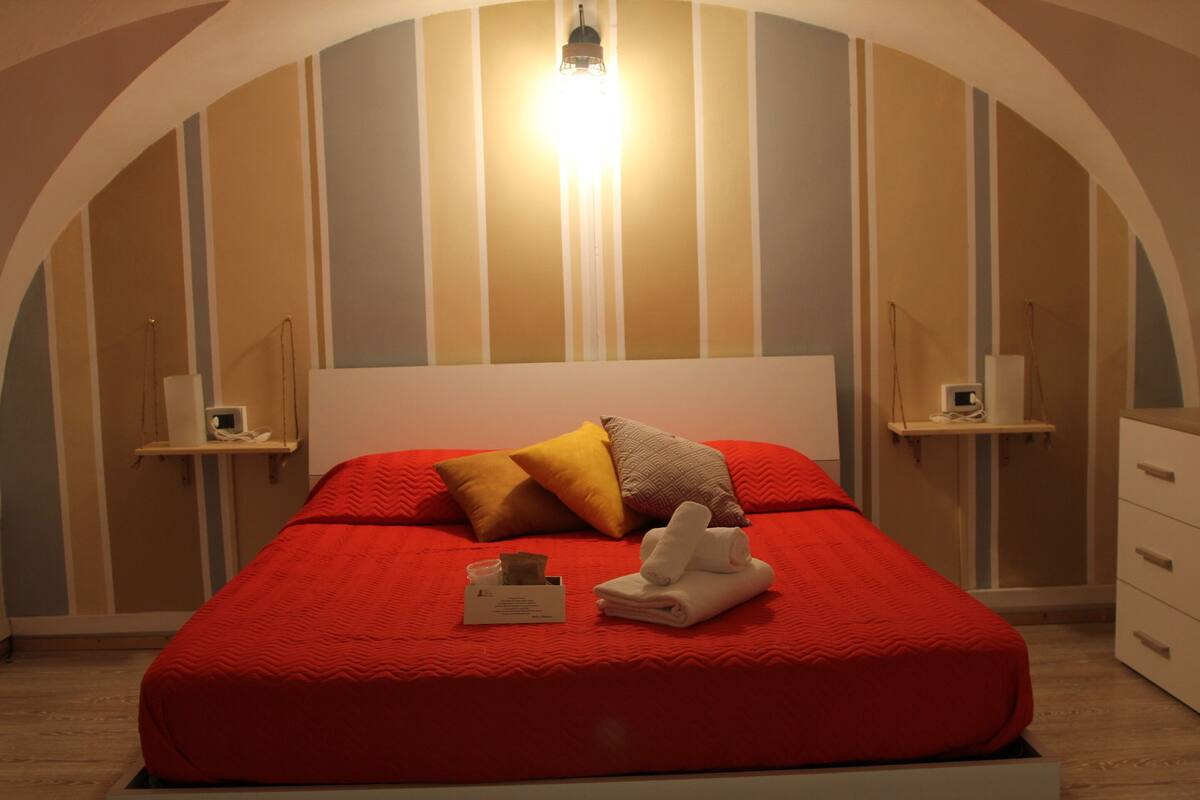 Un lit bien fait, recouvert d’une couverture rouge, avec plusieurs oreillers et une serviette pliée. Une table de chevet, sur laquelle se trouvent une lampe et une petite boîte, est placée à côté du lit. La pièce est décorée de motifs rayés sur les murs et possède un plafond incurvé.