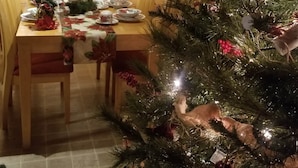 A dining area with a wooden table set for four, a Christmas tree with ornaments, and a window with blinds. - The Log Cabin on the Canoochee River (Evans County)