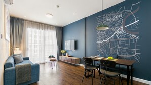 A modern living room with a blue sofa, wooden flooring, a dining area with a table and chairs, a TV on the wall, and a map mural. - Blue Fingers Saigon Royal - Tresor (Ho Chi Minh City)