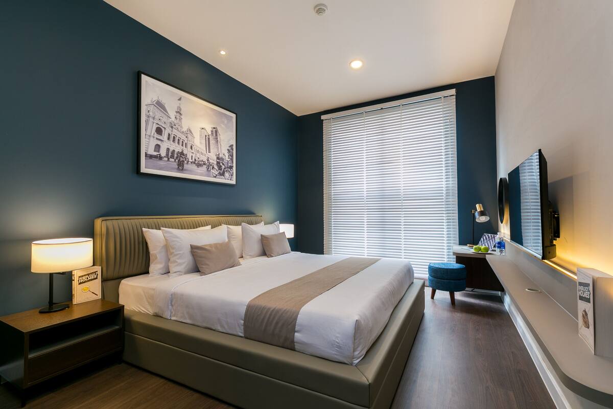A modern hotel room with a large bed, a bedside table with a lamp, a desk with a chair, a large window with blinds, and a framed picture on the wall.