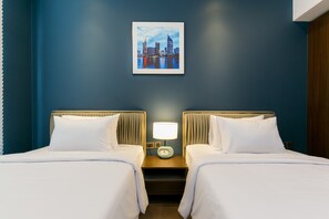 A hotel room with two beds, a nightstand with a lamp, and a cityscape painting on the wall.