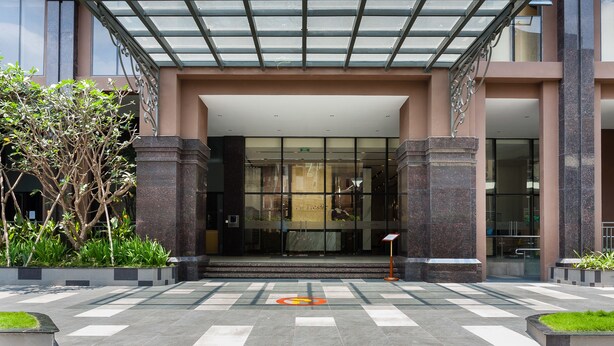 A modern building entrance with a glass canopy, a checkered floor pattern, and a small garden area. - Blue Fingers Saigon Royal - Tresor (Ho Chi Minh City)