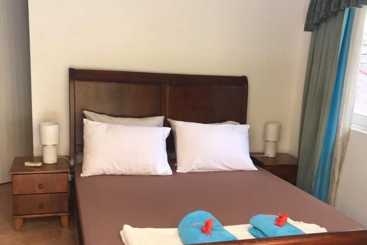 A single bed with a wooden headboard, two pillows, a nightstand with a lamp, and a window with curtains.