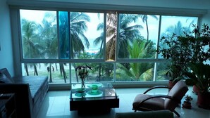 Ruime kamer met een groot raam dat uitzicht biedt op palmbomen en de oceaan. Er is een bank, een fauteuil en een klein tafeltje met decoratieve objecten.