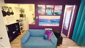 Een woonkamer met een blauwe bank, een kleine tafel en een groot raam met uitzicht op de oceaan. Er hangen keukengerei aan de muur en staat een plant op de vensterbank.