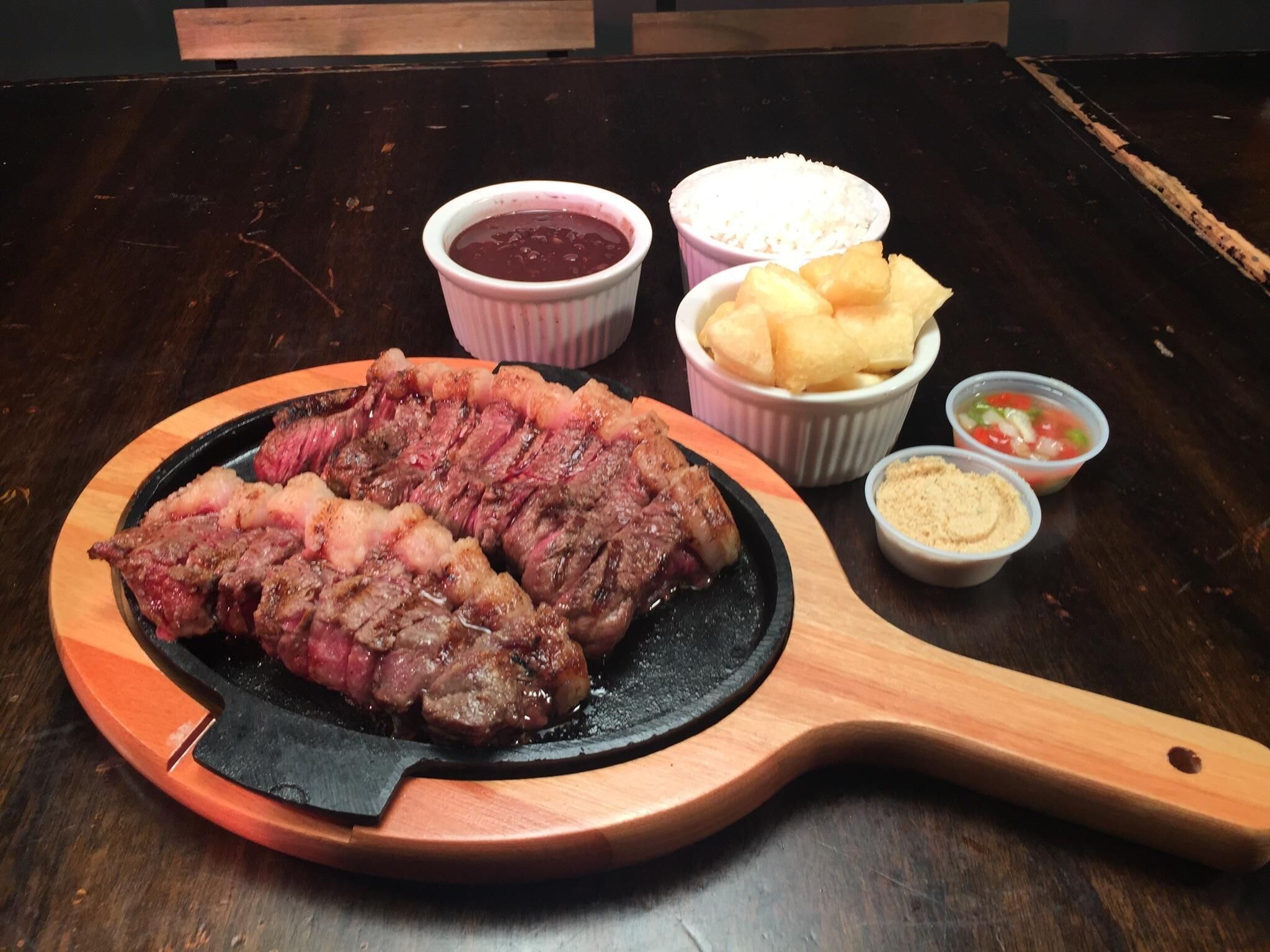 Un bistec sobre una tabla de madera con acompañamientos que incluyen un tazón de arroz, un tazón de salsa, un tazón de papas fritas y un pequeño tazón de salsa.