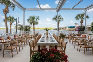 Un espace de restauration en plein air avec une longue table dressée pour un repas, entourée de chaises, et offrant une vue sur l’eau et un pont en arrière-plan.