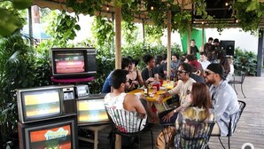Een groep mensen dineert in een openluchtrestaurant met meerdere televisies die verschillende programma's tonen.