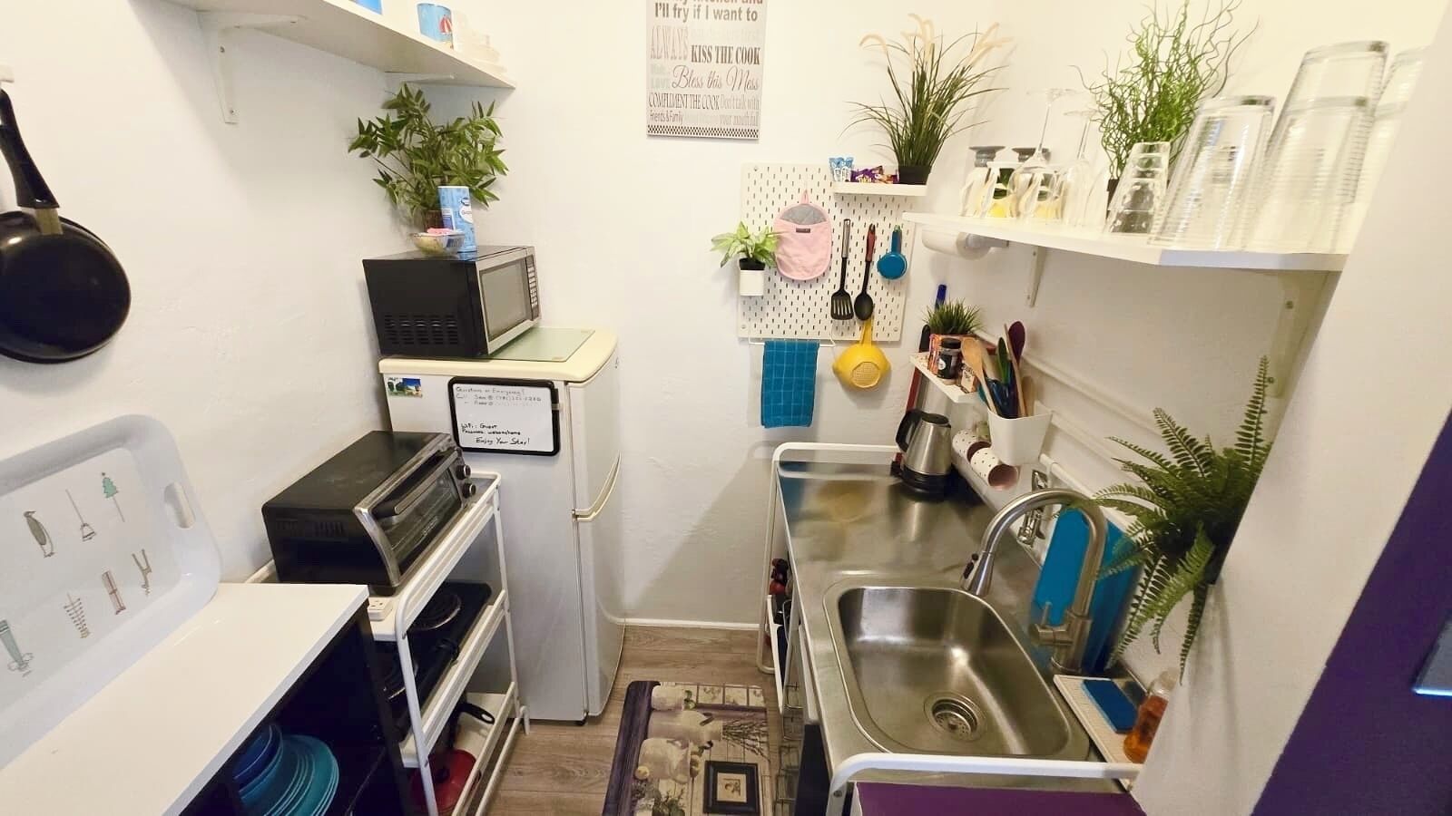 Une cuisine compacte équipée d’un four à micro-ondes, d’un grille-pain et d’un évier. On y trouve des étagères avec des plantes et des ustensiles de cuisine.