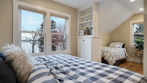 A bedroom with a large window, a bed with a striped bedspread, a white dresser, and a small plant. - Lake Home, salt water hot tub, sauna, boat rental (Forest Lake)