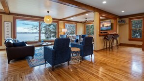 A spacious living room with a large window overlooking a lake, wooden flooring, and a cozy seating area with a sofa and chairs. - Lake Home, salt water hot tub, sauna, boat rental (Forest Lake)