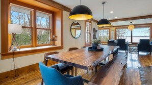 A spacious dining area with a long wooden table, black pendant lights, and large windows offering a view of the outdoors. - Lake Home, salt water hot tub, sauna, boat rental (Forest Lake)