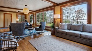 A spacious living room with a large window, a ceiling fan, and a grey sofa. - Lake Home, salt water hot tub, sauna, boat rental (Forest Lake)