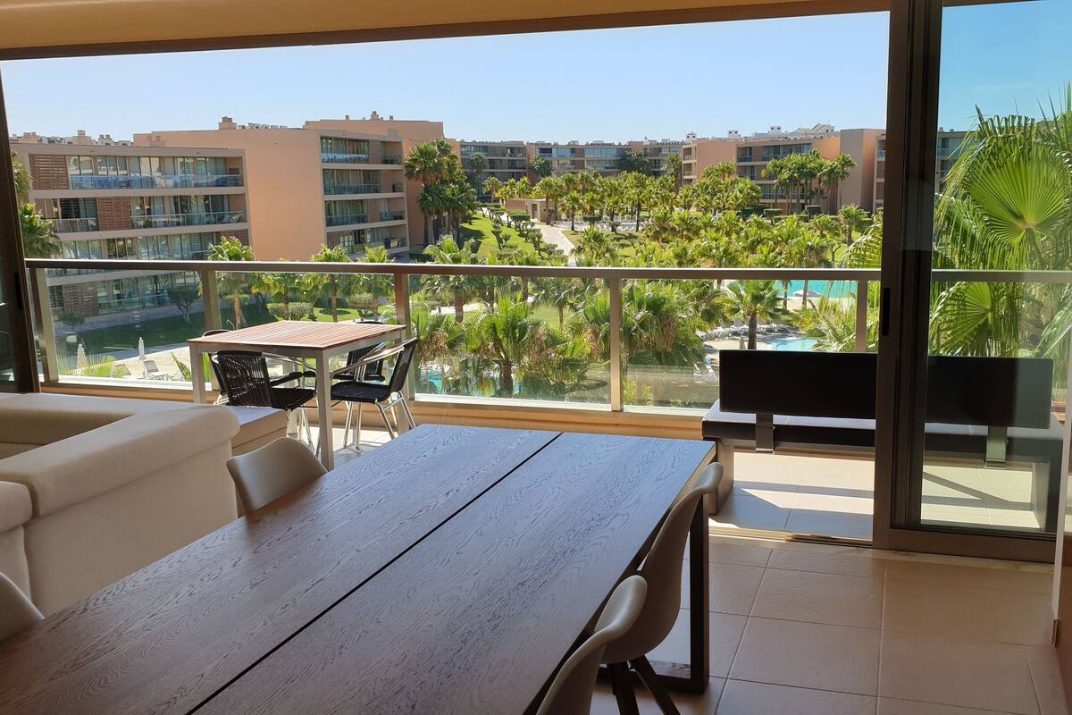A balcony with a wooden table and chairs, overlooking a pool and palm trees.