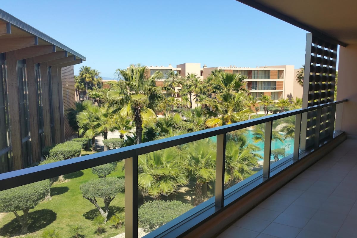 A balcony with a view of a swimming pool, palm trees, and modern buildings in the background.