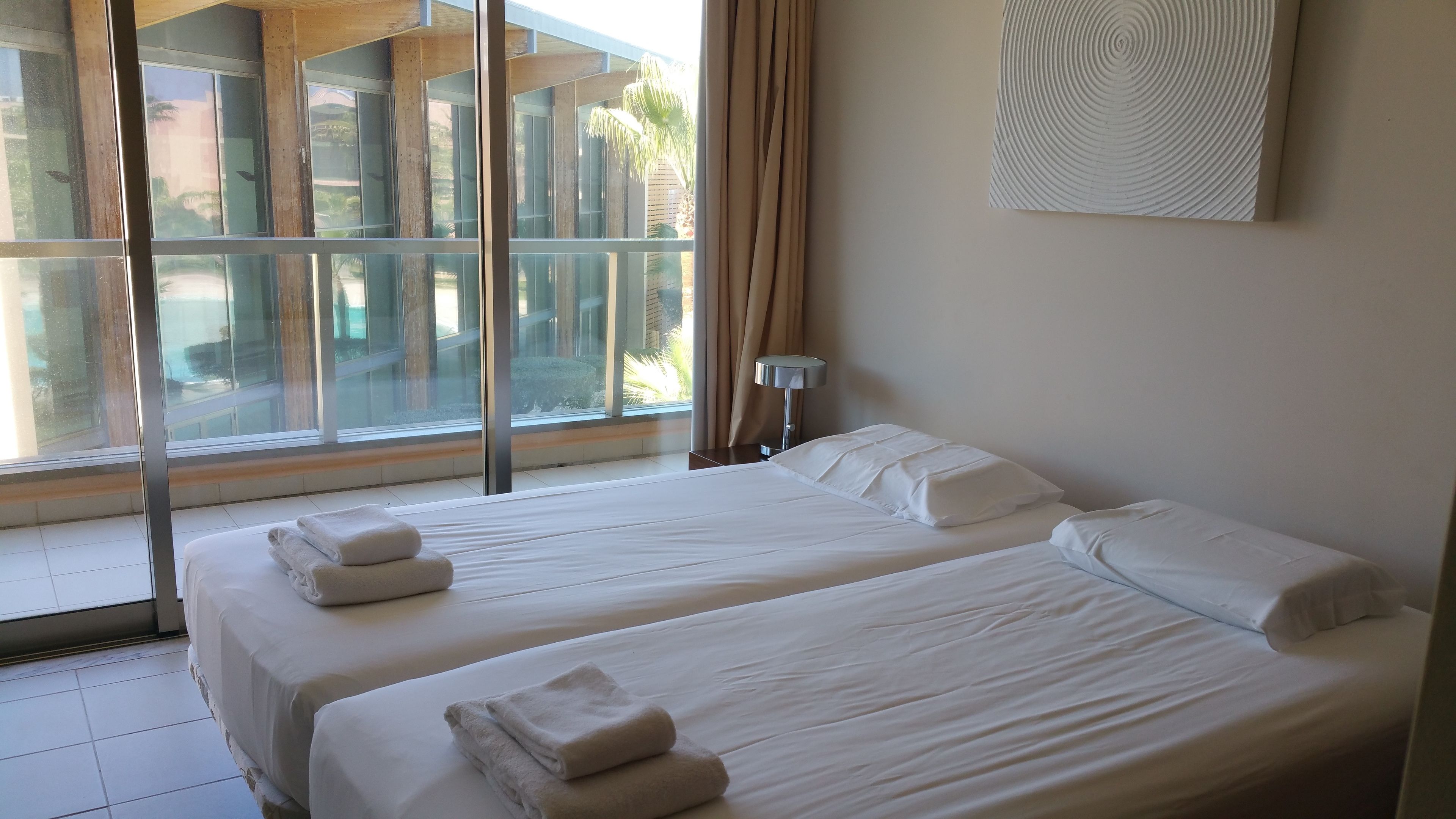 Een hotelkamer met twee bedden, een groot raam en uitzicht op een zwembad en palmbomen.